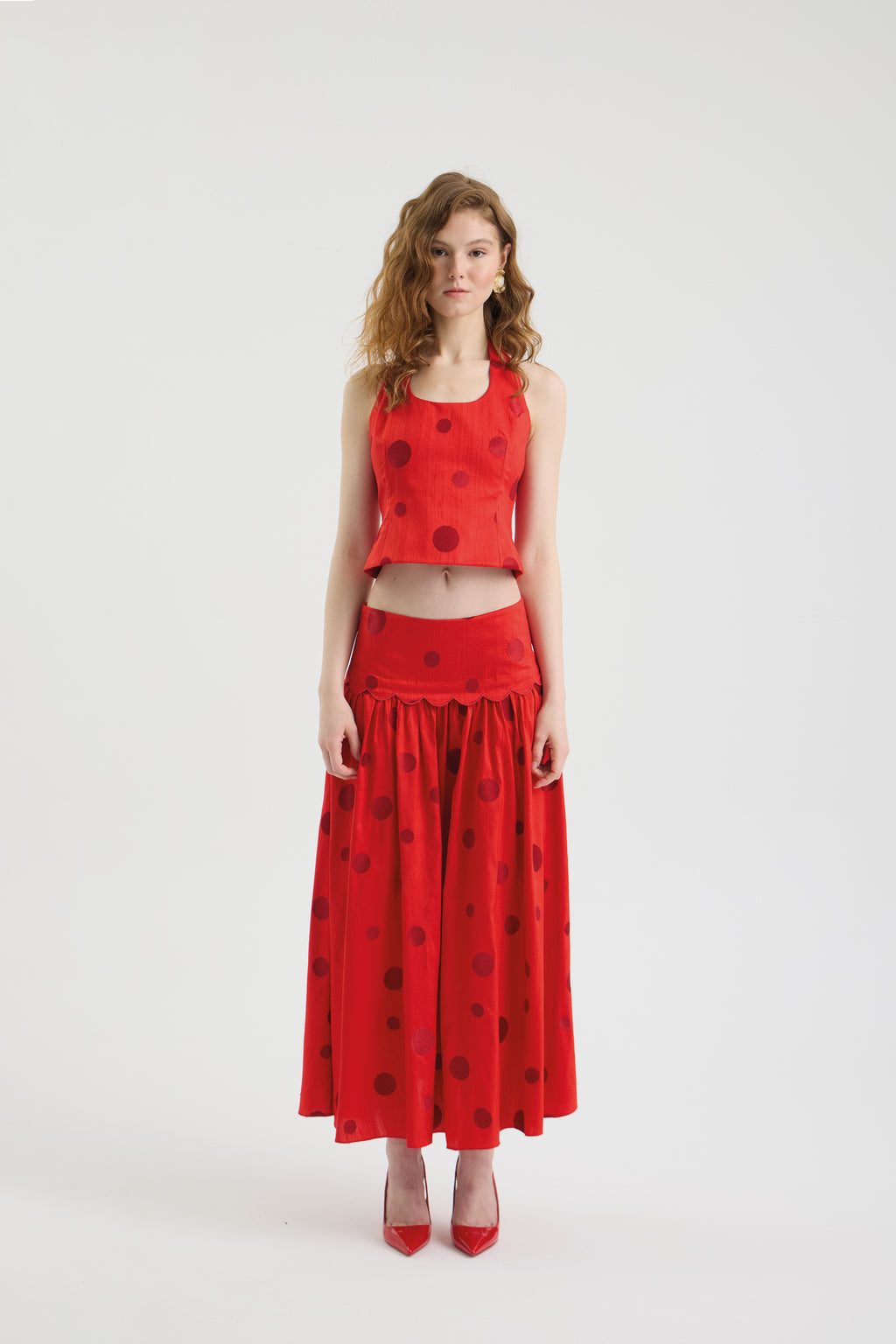 A woman stands against a white background wearing the POLKA FLAME TOP—a sleeveless silk shantung crop top—with a matching long red polka dot embroidered skirt and red high-heeled shoes.