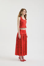 A woman stands sideways wearing the POLKA FLAME TOP, a red silk shantung crop top with large polka dot embroidery, paired with a matching skirt and red heels. She has long wavy hair and gold earrings, set against a white background.