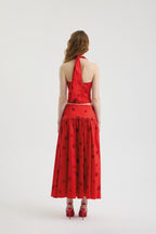 A woman with wavy brown hair faces away, wearing the POLKA FLAME TOP, a red silk shantung crop top with a halter neckline, paired with a matching red skirt featuring large polka dot embroidery and ruffles, plus red heels.