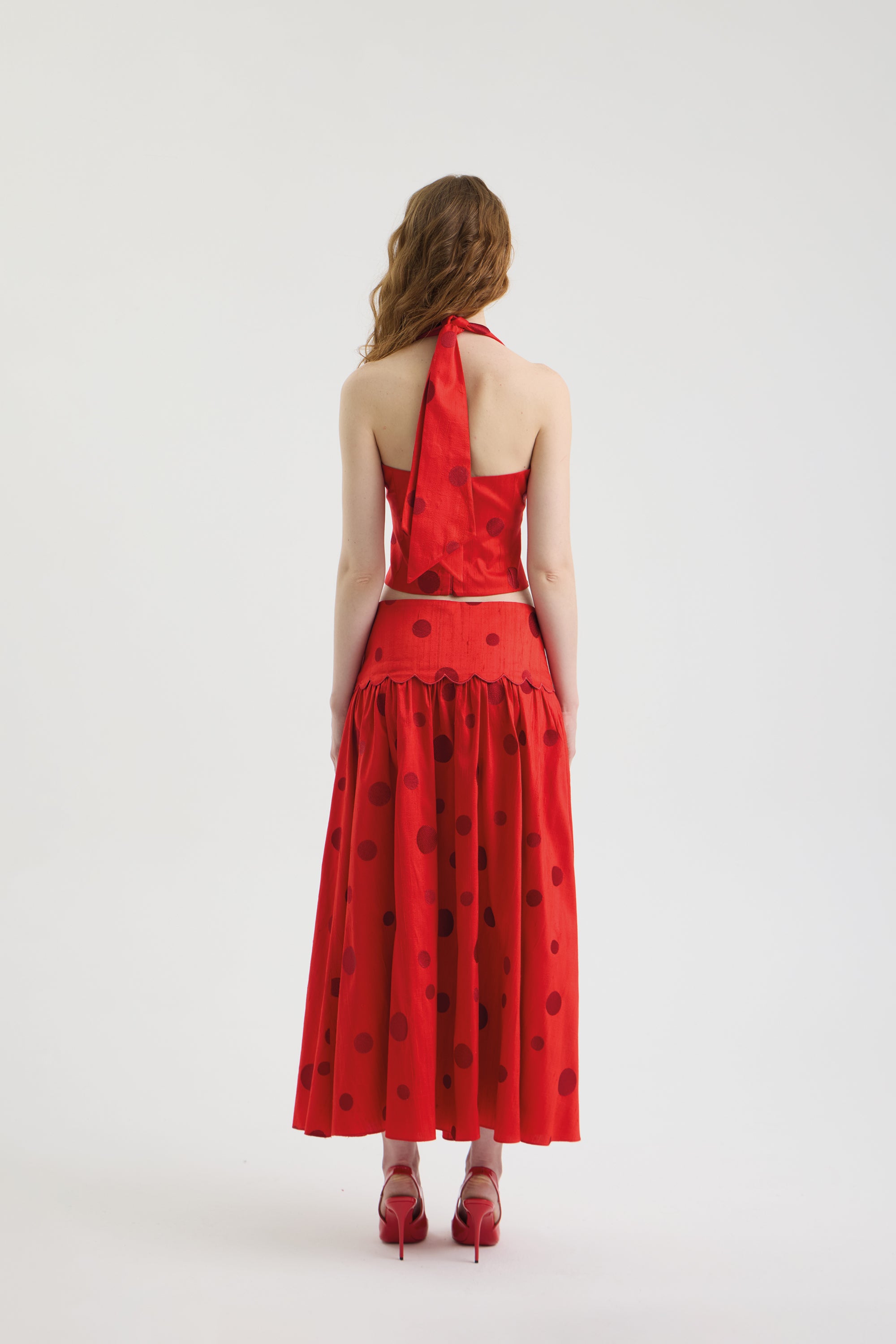 A woman with wavy brown hair faces away, wearing the POLKA FLAME TOP, a red silk shantung crop top with a halter neckline, paired with a matching red skirt featuring large polka dot embroidery and ruffles, plus red heels.