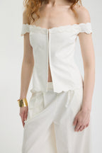 A woman wears the IVORY MUSE TOP, an off-shoulder corset with wave-shaped embroidery, paired with white wide-leg pants and a gold bracelet on her right wrist. Her wavy hair falls softly over her shoulders.