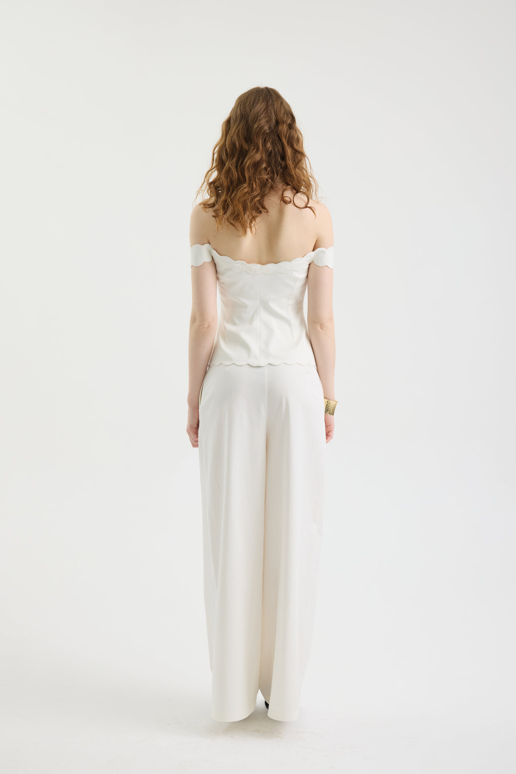A woman with wavy brown hair stands facing away in the IVORY MUSE TOP, an off-shoulder corset style, paired with wide-legged white pants featuring subtle wave embroidery against a plain white background.