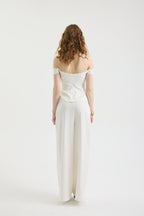 A woman with wavy brown hair stands facing away in the IVORY MUSE TOP, an off-shoulder corset style, paired with wide-legged white pants featuring subtle wave embroidery against a plain white background.