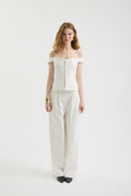 A woman with wavy, shoulder-length hair poses against a white background wearing the IVORY MUSE TOP—an off-shoulder corset with scalloped edges and wave embroidery—paired with loose white pants, black shoes, and a gold bracelet.