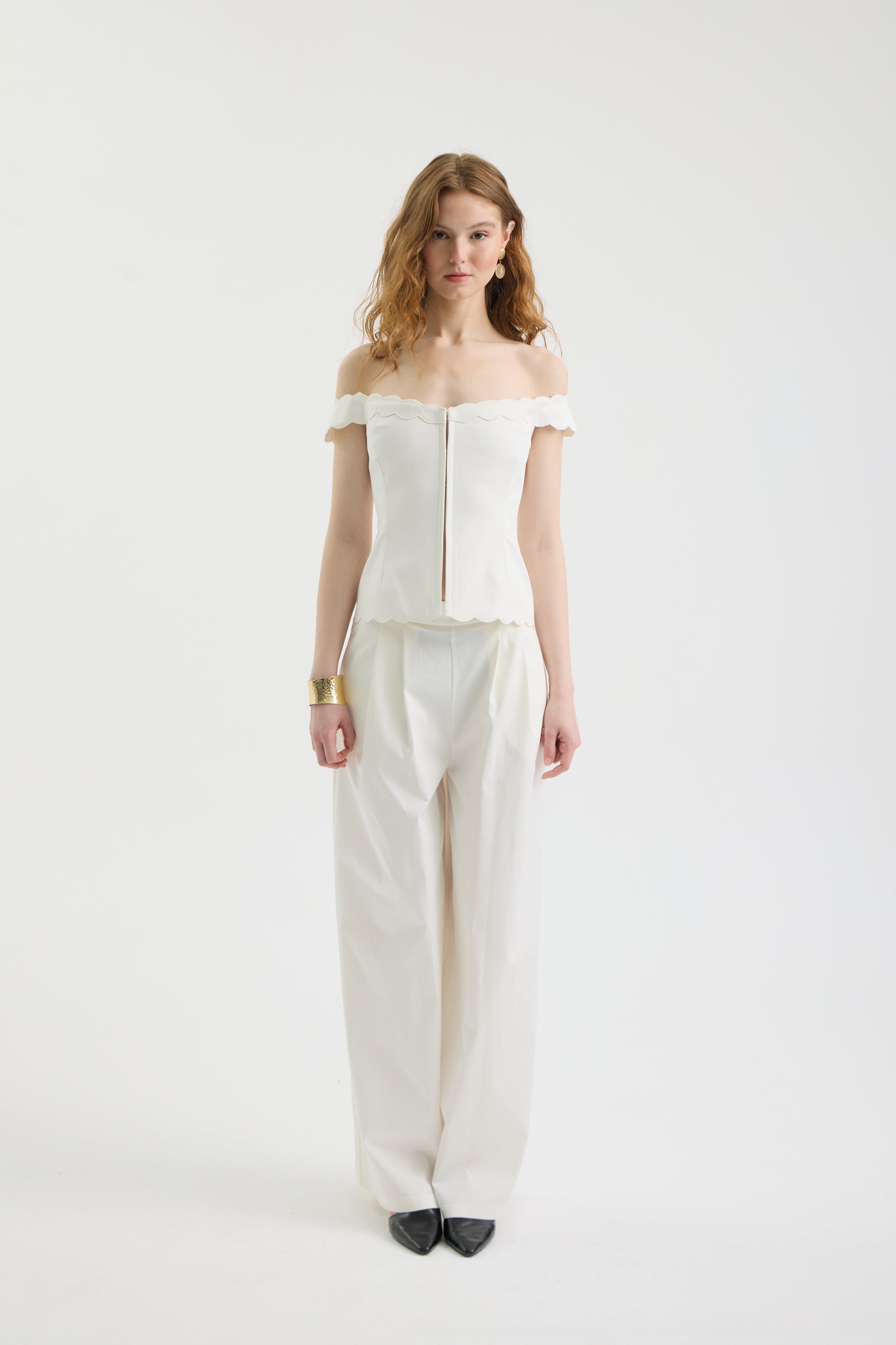 A woman with wavy, shoulder-length hair poses against a white background wearing the IVORY MUSE TOP—an off-shoulder corset with scalloped edges and wave embroidery—paired with loose white pants, black shoes, and a gold bracelet.