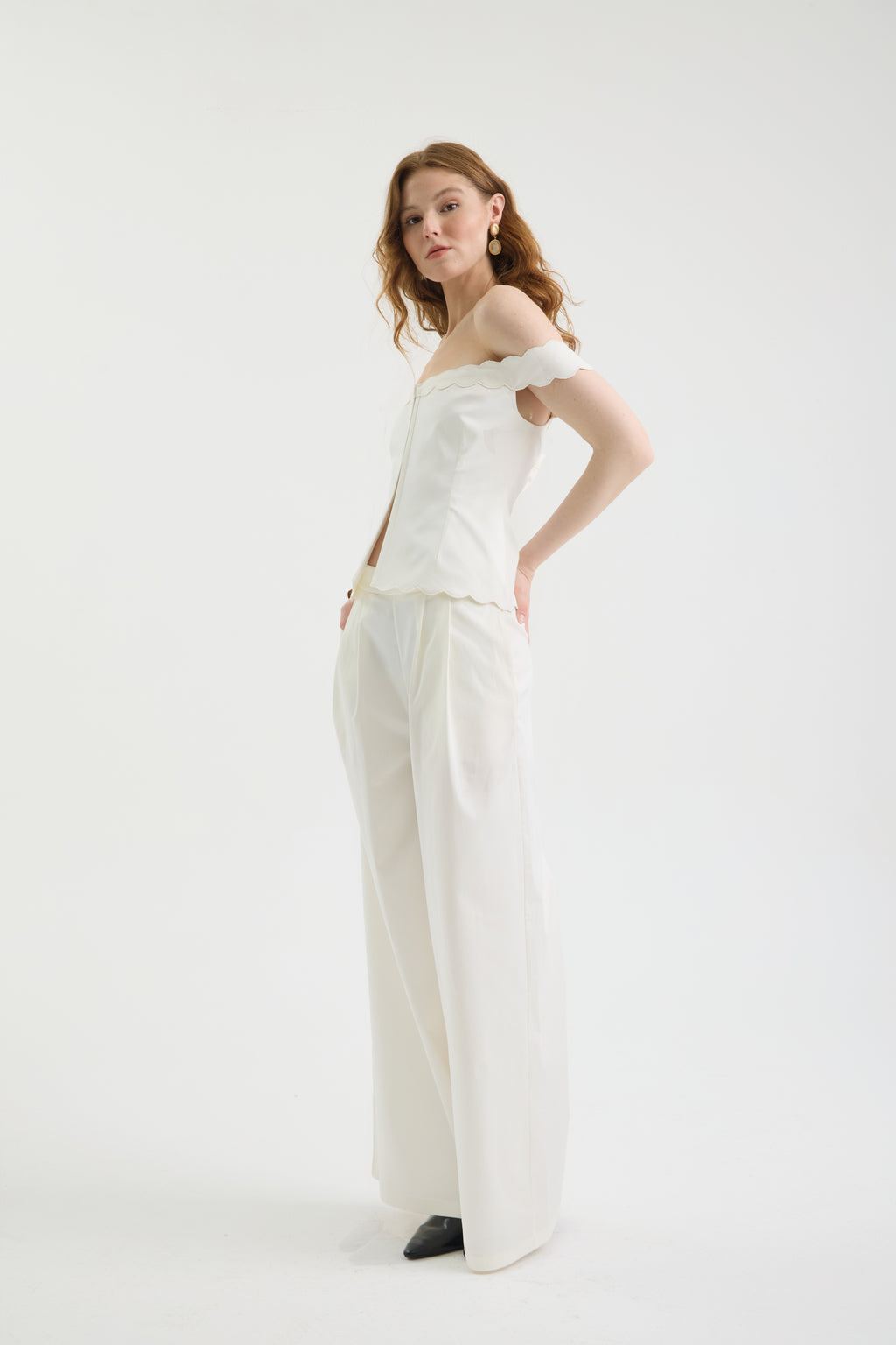 A woman with wavy hair stands confidently against a plain white background, wearing an off-shoulder white top and IVORY FLOW PANTS, styled with black shoes and gold earrings. One hand rests on her hip.