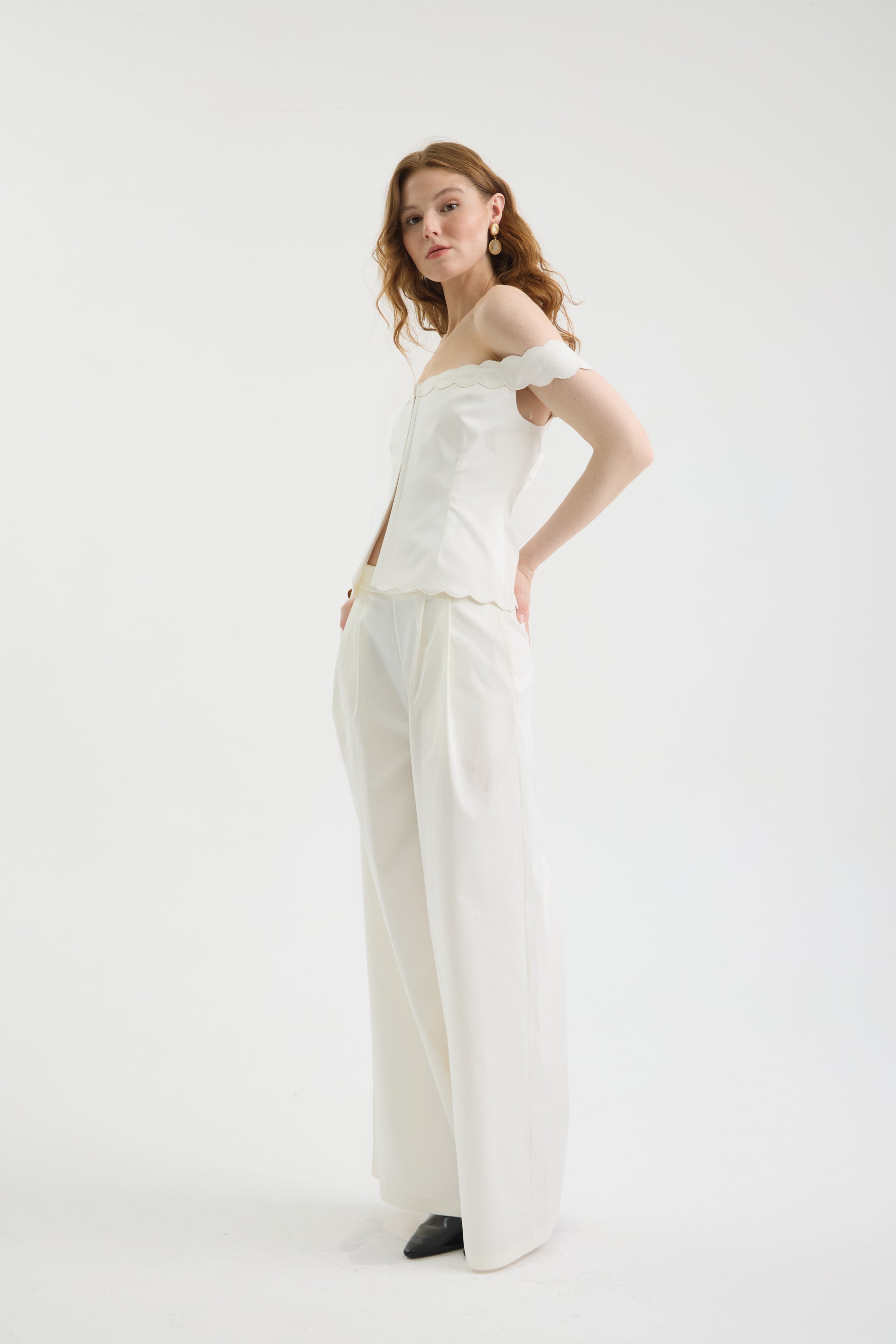 A woman with wavy hair stands confidently against a plain white background, wearing an off-shoulder white top and IVORY FLOW PANTS, styled with black shoes and gold earrings. One hand rests on her hip.