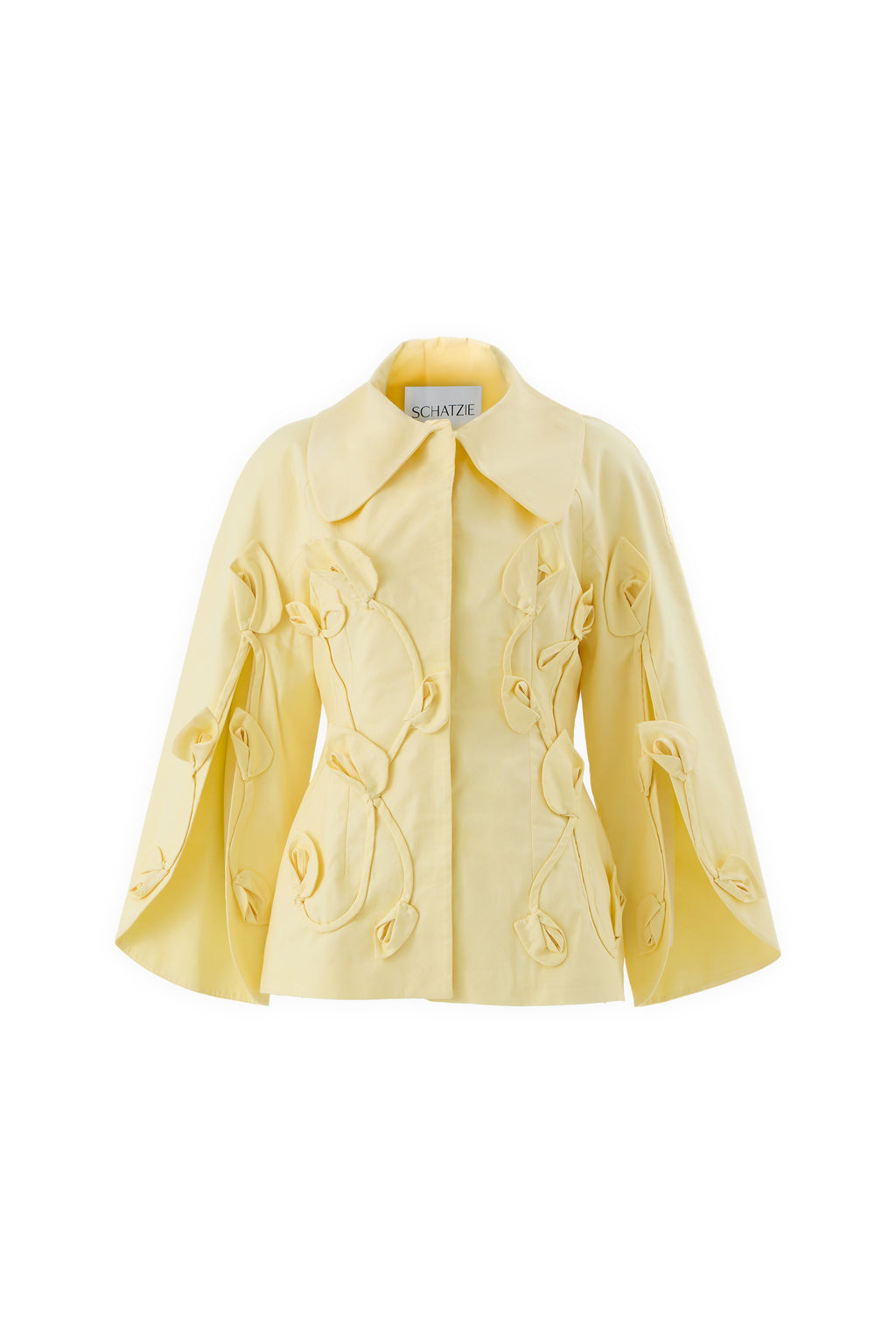 The LUMIÈRE BLOOM jacket features a pale yellow tailored design with a wide collar, cape-like sleeves, and handcrafted tulip appliqués. The label inside the collar reads SCHATZI.